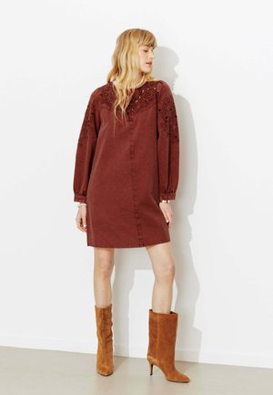 Femme portant une robe à manches longues couleur rouille avec des détails en dentelle et des bottes en daim à talons hauteur mollet de couleur beige, debout contre un mur blanc.