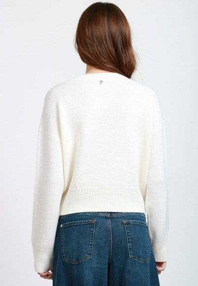Donna con lunghi capelli castani che indossa un maglione bianco a maglia e pantaloni di jeans blu a vita alta, di spalle rispetto alla fotocamera.
