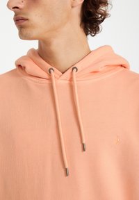 Haut à capuche de couleur pêche avec un cordon de serrage, des poignets côtelés et un petit logo brodé sur la poitrine. Texture lisse, design décontracté.