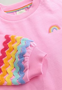 Rosa Sweatshirt mit einem gestickten Regenbogenmotiv und mehrfarbigen, rüschenden, gewellten Akzenten an einem Ärmel. Weicher Stoff.