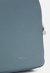 Light blue synthetic bag with a smooth texture, featuring a metallic zipper and a round logo charm. Brand name "ANNA FIELD" displayed prominently.