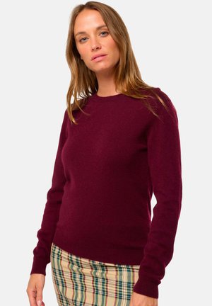 Pull en laine bordeaux à col rond et manches longues, associé à une jupe à carreaux avec des tons crème, vert et rouge.