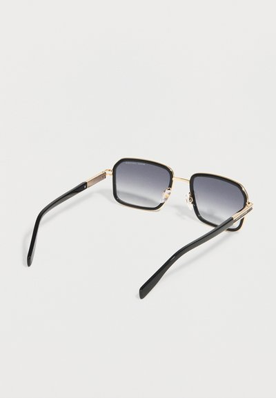 Γυαλιά ηλίου τετράγωνου σχήματος με πλαίσια σε μαύρο και χρυσό χρώμα και σκοτεινούς φακούς με διαβάθμιση, τοποθετημένα πάνω σε λευκή επιφάνεια.