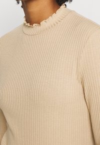 Beige Ripp-Top mit einem hohen, rüschigen Ausschnitt. Der Stoff ist glatt mit subtiler Textur und zeigt vertikale Streifen für zusätzlichen Detail.