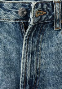 Svijetloplave traperice s patentnim zatvaračem, srebrnim gumbom i kontrastnim narančastim šavovima duž šavova i džepova.