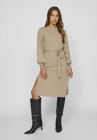 Abito in maglia beige con maniche lunghe, vita cintrata, apertura laterale e vestibilità rilassata. Abbinato a stivali neri alti con punta appuntita.