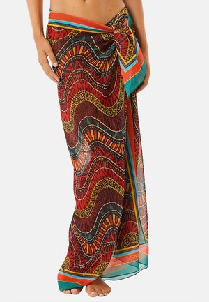 Donna che indossa un sarong colorato con motivi, legato in vita, con disegni ondulati rossi, gialli, neri e blu, in piedi a piedi nudi.
