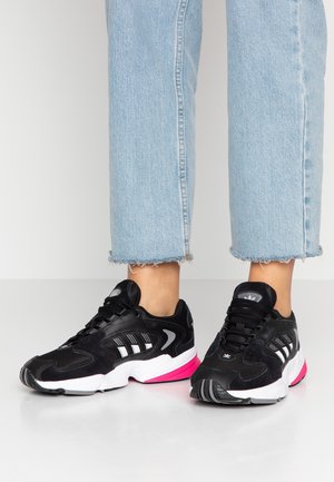 Person iført sorte og hvide sneakers med pink accenter og lyseblå korte jeans, der står på en hvid gulv.