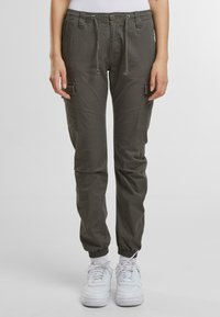 Olive zelené cargo nohavice, vyrobené z ľahkého materiálu, s elastickými manžetami, sťahovacím pásom a dvoma bočnými vreckami. Na nohách sú vidieť biele tenisky.