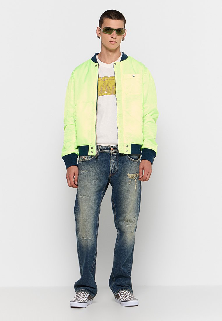 Veste bomber verte fluo avec des accents bleu marine, t-shirt blanc avec un graphique jaune, jeans bleus déchirés, et chaussures slip-on à carreaux.