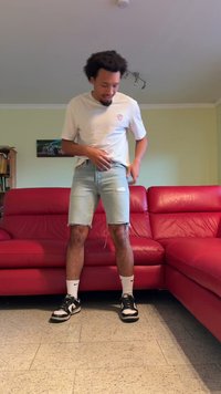 Ανοιχτό μπλε τζιν σορτς με φθαρμένα τελειώματα, φορεμένο με λευκό t-shirt και μαύρα αθλητικά παπούτσια, που στέκεται σε υφή δαπέδου δίπλα σε έναν κόκκινο δερμάτινο καναπέ.