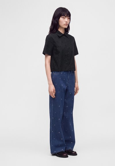 Femme aux longs cheveux foncés portant une chemise noire à manches courtes boutonnée et un jean bleu large avec de petits motifs blancs, debout de profil.