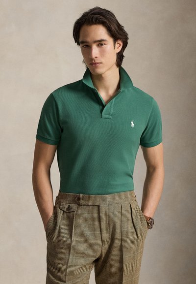 Polo vert à texture côtelée avec un col classique, associé à un pantalon marron à carreaux avec des plis et des poches latérales.