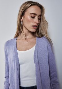 Lavendel kofta med öppen front, mjuk textur, långa ärmar och en avslappnad passform, lager på lager över en vit tank topp. Hudton neutral bakgrund.