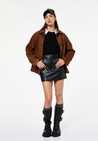 Vrouw in een bruine jas, zwarte trui, gestreept shirt, zwarte leren mini-rok, zwarte gespschoenen en een zwarte pet, staand met haar handen op haar heupen.