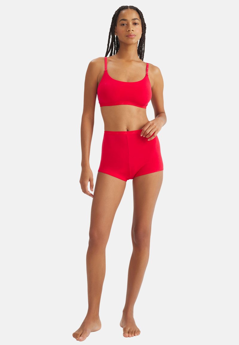 Kobieta stojąca boso, ubrana w dopasowany, jaskrawoczerwony biustonosz i szorty z wysokim stanem na jednolitym białym tle.