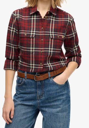 Kvinde iført en rød og sort ternet skjorte med knapper og opkørte ærmer, blå jeans og et brunt læderbælte med en sølvspænde.