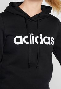 Felpa nera con un prominente logo bianco "adidas". Presenta un cappuccio con cordino e una texture morbida, adatta per un abbigliamento casual.
