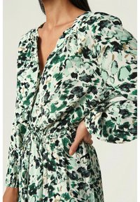 Vestido floral con un estampado abstracto en verde y negro, mangas largas abullonadas, escote en V y una cintura elástica con un detalle de cordón.