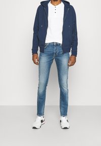 Granatowa bluza z kapturem na zamek, biała koszula henley, niebieskie dżinsy i białe sneakersy. Bluza ma boczne kieszenie i haftowane logo na rękawie.