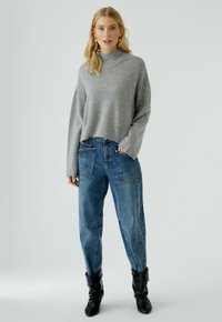 Lichtgrijze trui met een hoge nek en lange mouwen, gecombineerd met hoge taille blauwe jeans met patch zakken en contrasterende stiksels. Zwarte enkellaarzen.