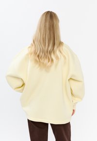 Przesadny żółty sweatshirt o teksturowanym wykończeniu, z opadłymi ramionami i ściągaczami, zestawiony z brązowymi spodniami z korduroju.