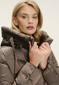 Smeđa puffer jakna s visokim ovratnikom, sjajnom površinom i kvadratnim uzorkom. Sadrži vezice i dugmadi za zatvaranje na manžetama.