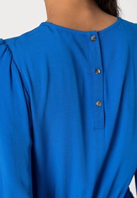 Sininen mekko, jossa on sileä pinta, pyöreä kaula-aukko, lyhyet puffihihat ja kolme koristepainiketta takana.