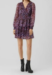 Mujer vestida con un vestido de manga larga a rayas de cebra morado y marrón, combinado con botas de tobillo de plataforma negras, de pie contra un fondo sencillo.