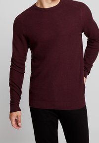 Mann in einem enganliegenden, langärmligen weinroten Strukturstrickpullover mit gerippten Bündchen und Saum, kombiniert mit schwarzen Hosen.