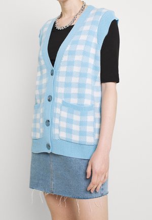 Personne portant un gilet boutonné à carreaux bleus et blancs sur un haut noir, associé à une jupe en jean bleue à ourlet effiloché.