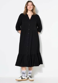 Zwart button-up jurk met een gelaagde zoom, 3/4 mouwen en een gestructureerde stof. Gecombineerd met witte patroon sokken en zwarte sneakers.
