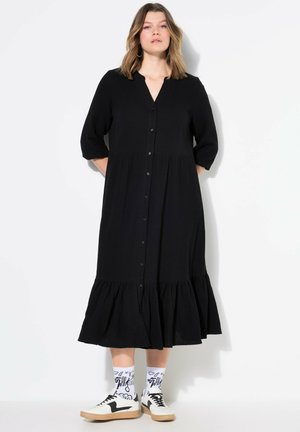 Zwart button-up jurk met een gelaagde zoom, 3/4 mouwen en een gestructureerde stof. Gecombineerd met witte patroon sokken en zwarte sneakers.