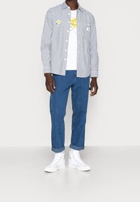 Camisa de cuadros en azul y blanco, llevada sobre una camiseta blanca con un gráfico de carita sonriente, combinada con pantalones de mezclilla azules y zapatillas blancas.