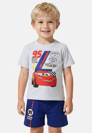 Ragazzo giovane sorridente, indossa una maglietta grigia e pantaloncini blu con disegni di auto da corsa rosse e a scacchi e il numero 95.