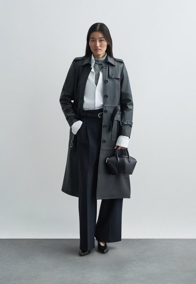 Manteau trench en cuir noir avec des boutons, des revers et des poches, porté sur une chemise blanche à col et un pantalon large sombre ; tenant un petit sac noir.
