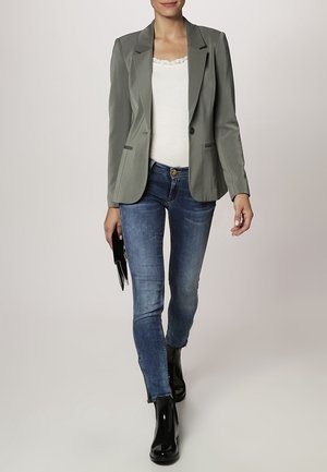 Blazer gris avec revers crantés, bouton unique, porté avec un débardeur blanc, un jean skinny bleu et des bottines noires, tenant une pochette.