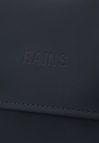 Sac imperméable bleu marine avec une texture mate lisse. Présente un logo "RAINS" en relief et un détail de bordure soigneusement cousu.
