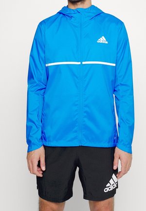 Mężczyzna w jasnoniebieskiej kurtce Adidas na suwak z kapturem i czarnych szortach Adidas, stojący na jednolitym białym tle.