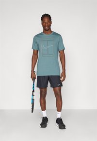 Τιρκουάζ βαμβακερό t-shirt με λευκό λογότυπο Nike και σχέδιο περιγραμμάτων σε ορθογώνια, συνδυασμένο με μαύρες βερμούδες, λευκές κάλτσες και μαύρα αθλητικά παπούτσια.