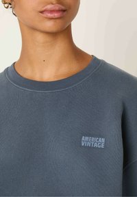 Tamno plavi dukserica od mekog materijala, s rebrastim okruglim izrezom i malim otisnutim logotipom "AMERICAN VINTAGE" na prsima.
