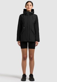 Noor naine musta kapuutsiga jope, millel on kardinapaelad, mustad lühikesed püksid ja mustad tossud, seisab sirgega heleda tausta ees.