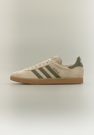 Zapatilla Adidas Gazelle de ante beige con rayas verdes, diseño con cordones y suela de goma, mostrada de perfil sobre un fondo neutro.