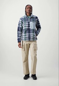 Person iført en blå, grøn og hvid ternet fleecepullover med beige cargo bukser og sorte sko, stående imod en ensartet lys baggrund.