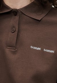 Chemise marron à col avec deux boutons et texte blanc « SAMSØE SAMSØE » brodé sur la poitrine.