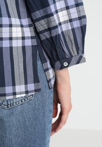 Camicia a quadri blu e viola con maniche lunghe e voluminose. Presenta un polsino con bottone e spacco laterale. Indossata con jeans di tessuto medio.