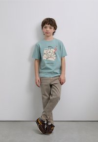 T-shirt en coton bleu clair avec un imprimé graphique de tête de mort et os croisés, associé à un pantalon beige et des chaussures de sport marron.