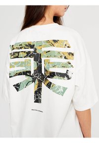 Bílý oversized tričko s velkým, prasklým, abstraktním grafickým potiskem v černé, zelené a zlaté barvě na zadní straně, s textem "MULTIPLY APPAREL".