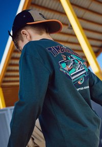Jeune personne portant un sweat-shirt Timberland vert foncé et une casquette noire et beige, se tenant sous une structure métallique jaune avec un ciel bleu en arrière-plan.
