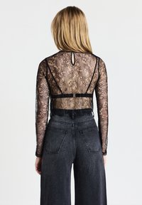 Schwarzes Spitzenshirt mit langen Ärmeln und einem Schlüsselaugen-Design auf der Rückseite, kombiniert mit hoch taillierten Jeans. Die Spitze zeigt florale Muster.